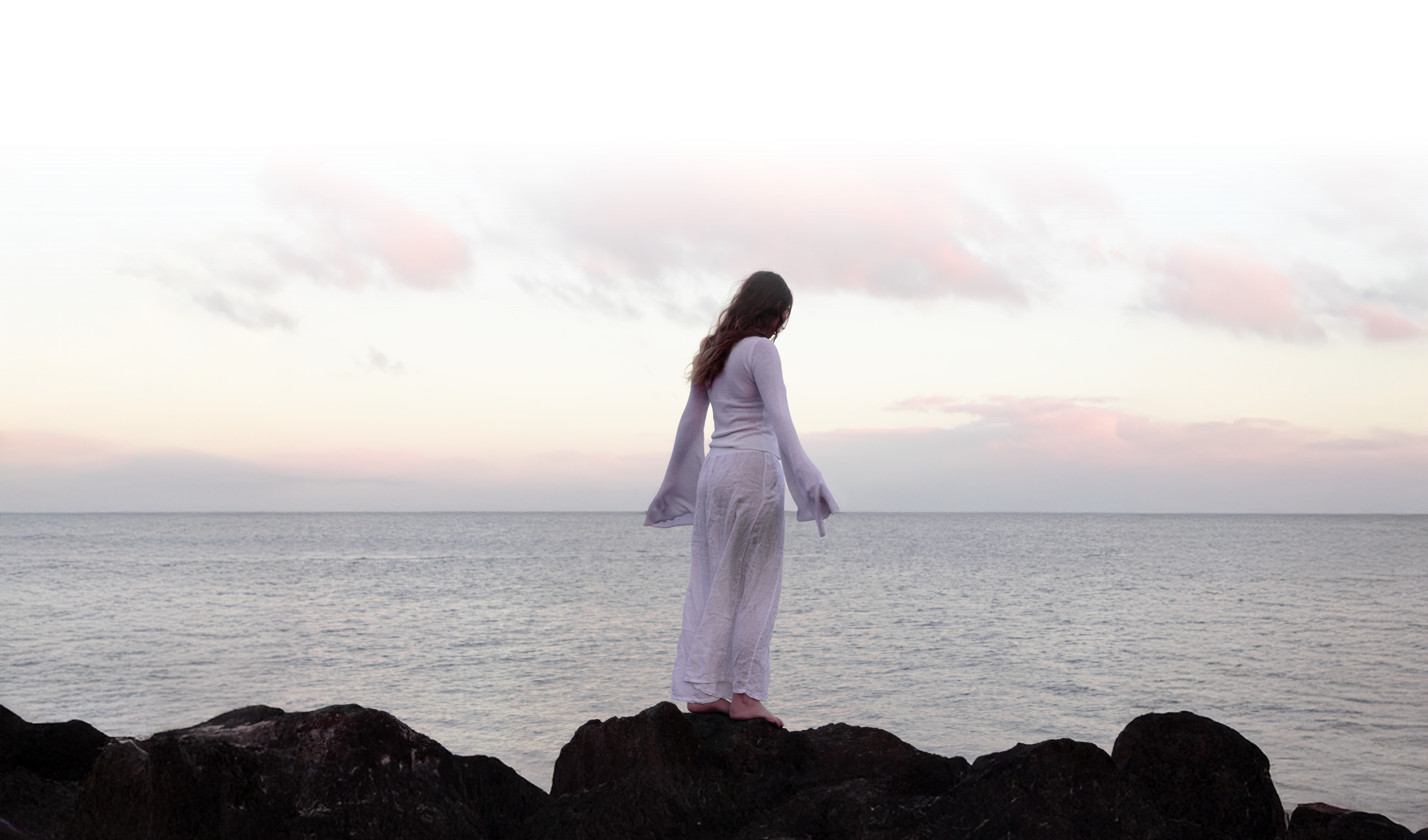 Person in a white outfit standing on rocks overlooking a vast ocean at sunset.
